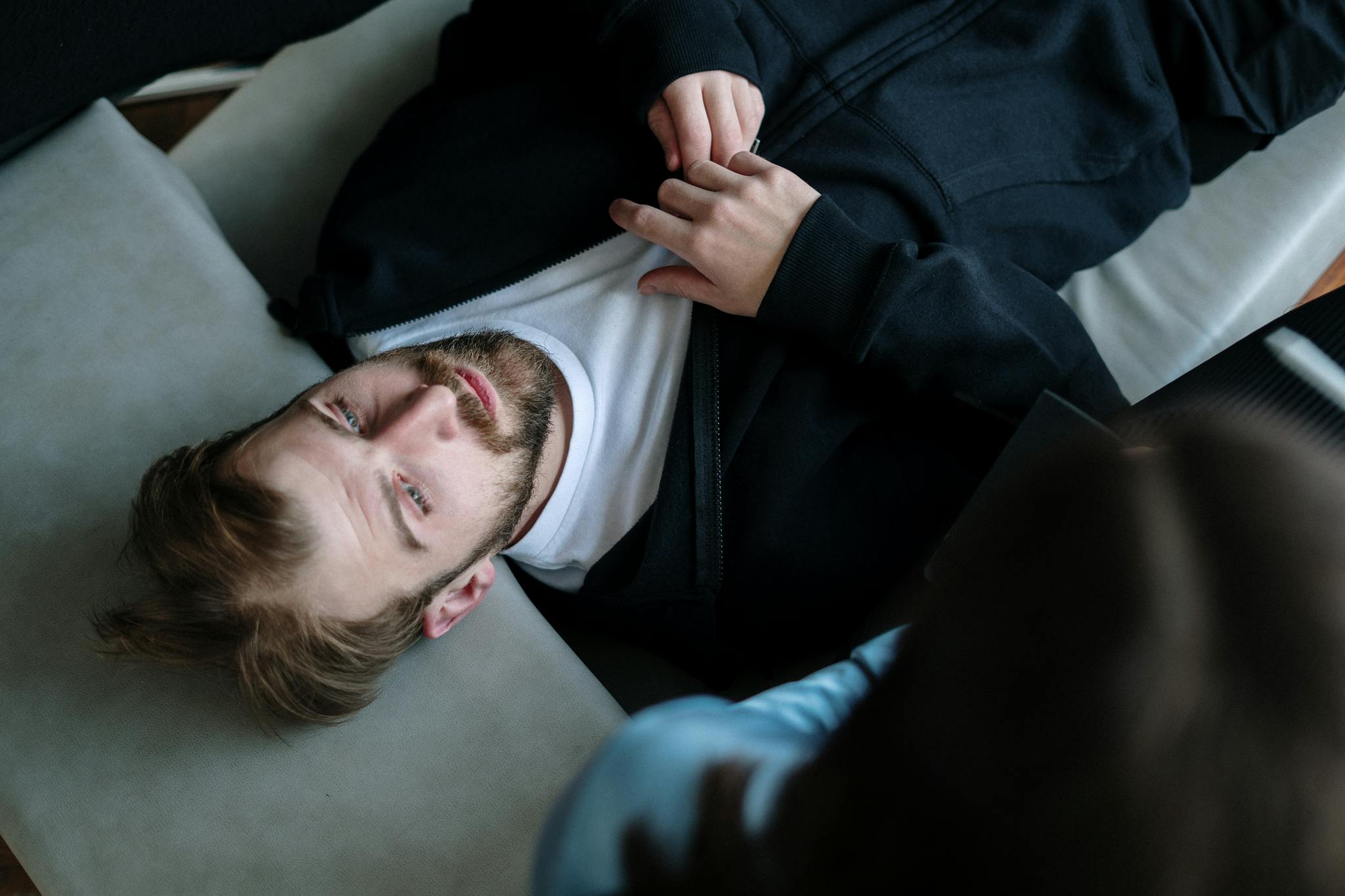 A bearded man lying down during a therapy session indoors, expressing emotions.
